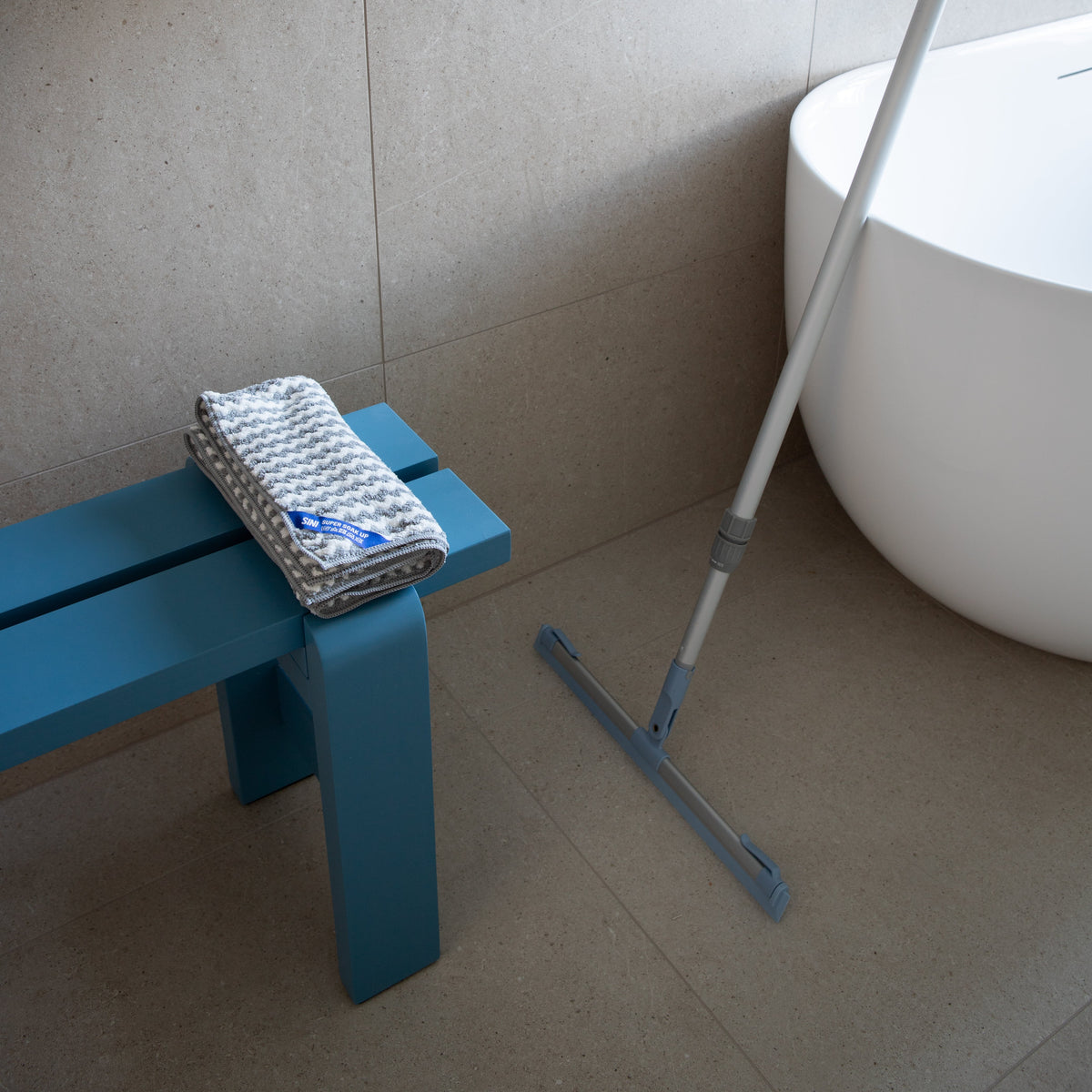 A blue wooden bench with a folded patterned towel sits next to a white freestanding bathtub in a tiled bathroom, with a long-handled squeegee leaning against the tub wall.