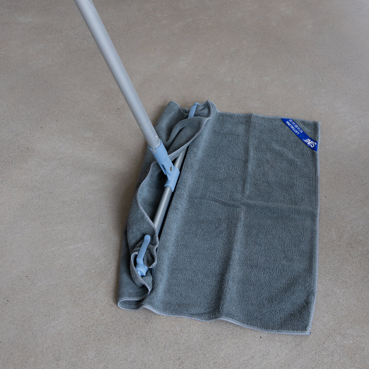 A gray microfiber cleaning cloth attached to a silver and blue mop handle, lying on a beige carpeted floor with wooden chair legs visible in the background.