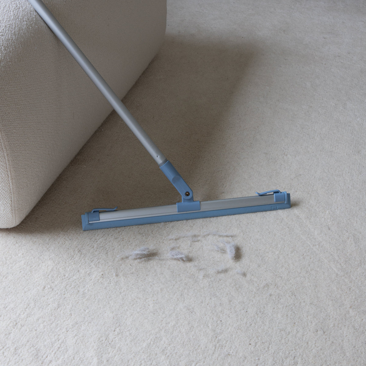 A blue and silver floor squeegee with a long handle resting on a white shag rug in a living room setting.